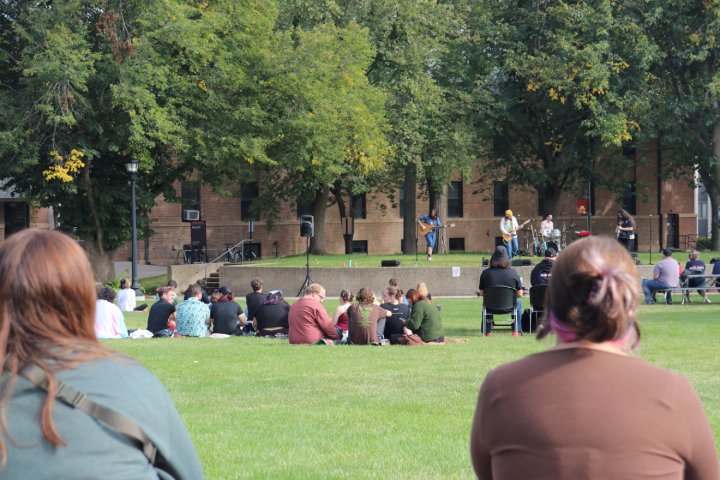 People sitting on the grass and in chairs, watching a live outdoor concert on a campus lawn. A band performs on a small stage in front of a backdrop of trees and a brick building.